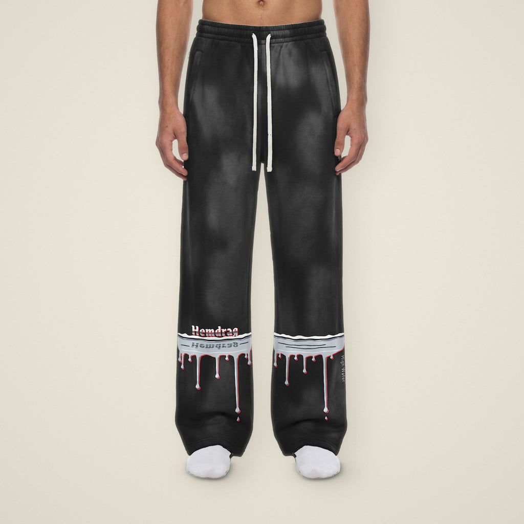Digital preview: front view of a model in black sunfade High Water wide-leg sweatpants. A chrome flood line with drips wraps both lower legs; on the right leg Hemdrag stands on the waterline with its reflection in the chrome, and High Water runs down the outer seam of the left leg. The print and the black color were added digitally to a model photo.