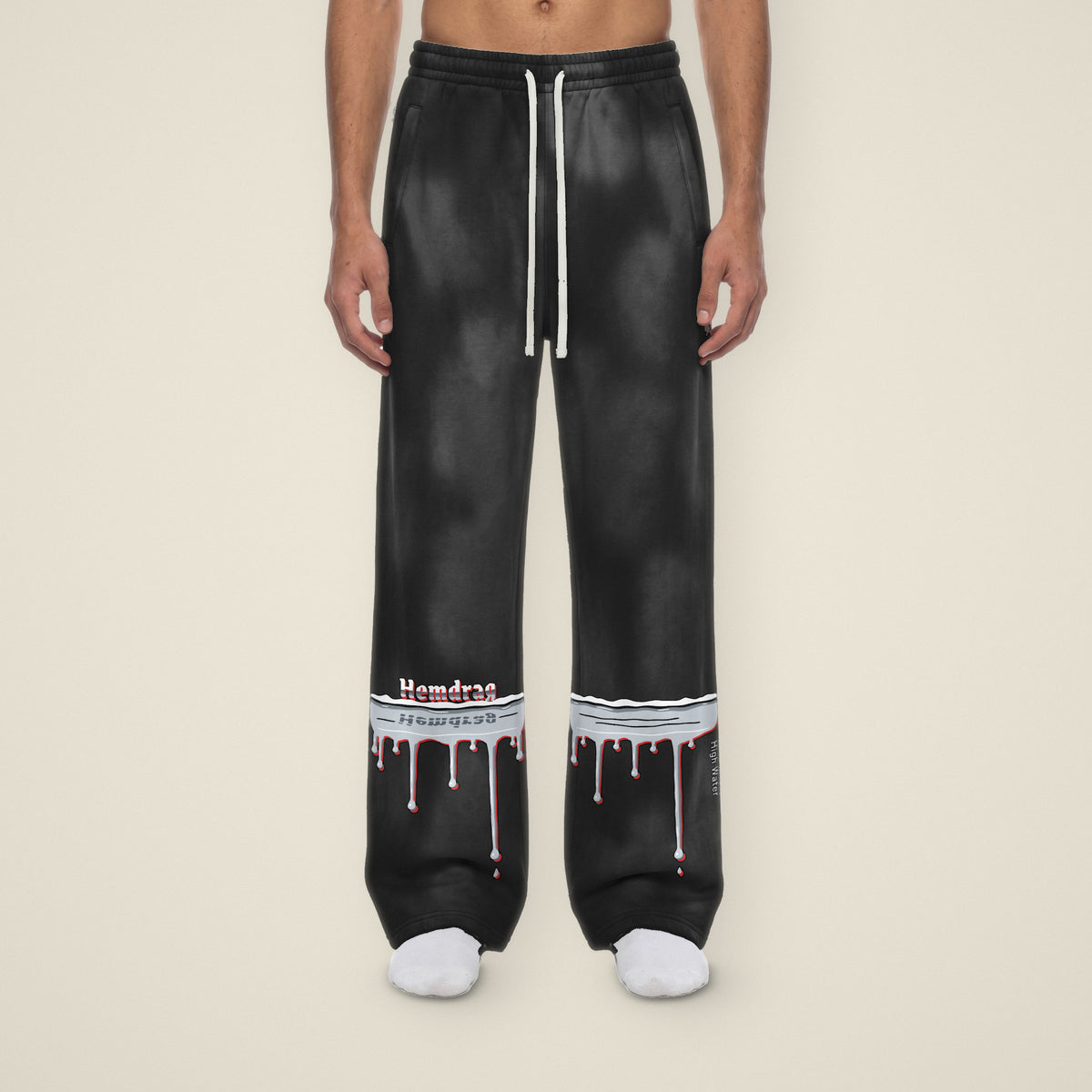 Digital preview: front view of a model in black sunfade High Water wide-leg sweatpants. A chrome flood line with drips wraps both lower legs; on the right leg Hemdrag stands on the waterline with its reflection in the chrome, and High Water runs down the outer seam of the left leg. The print and the black color were added digitally to a model photo.