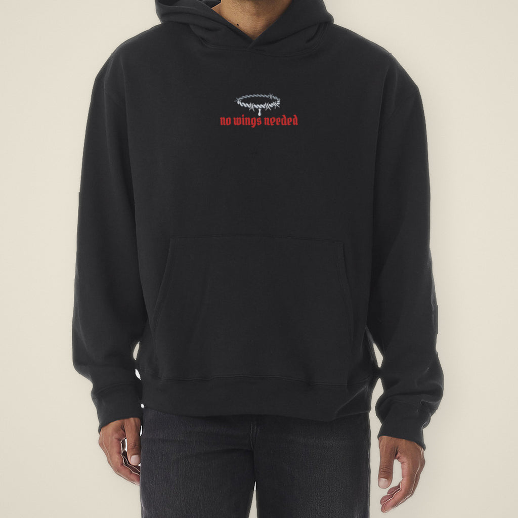 Digital preview: front of the black No Wings Needed hoodie on a model, cropped at the chin: a small chrome barbed-wire halo with one drip above "no wings needed" in red blackletter. Shown on a similar black hoodie used as a stand-in, from the same shoot as the back view.