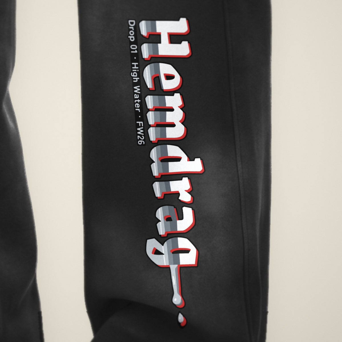 Digital preview: close-up of the whole leg print: the chrome Hemdrag letters with a red offset from the H to the g, which melts into a drip and a falling drop, beside the small line "Drop 01 · High Water · FW26". The print and fabric texture are simulated.