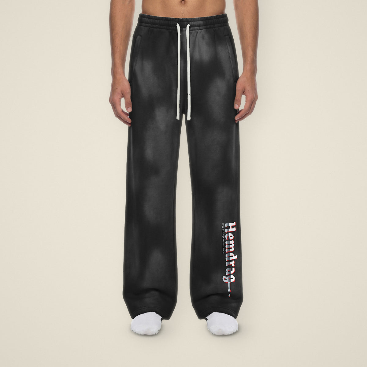 Digital preview: front view of a model in the Hemdrag sweatpants, showing the one-sided design: Hemdrag down the left lower leg, the right leg left clean. The print and the black color were added digitally to a model photo.